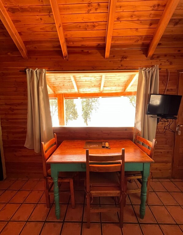 A wooden cabin with a dining table and chairs, a TV mounted on the wall, and a window with curtains.