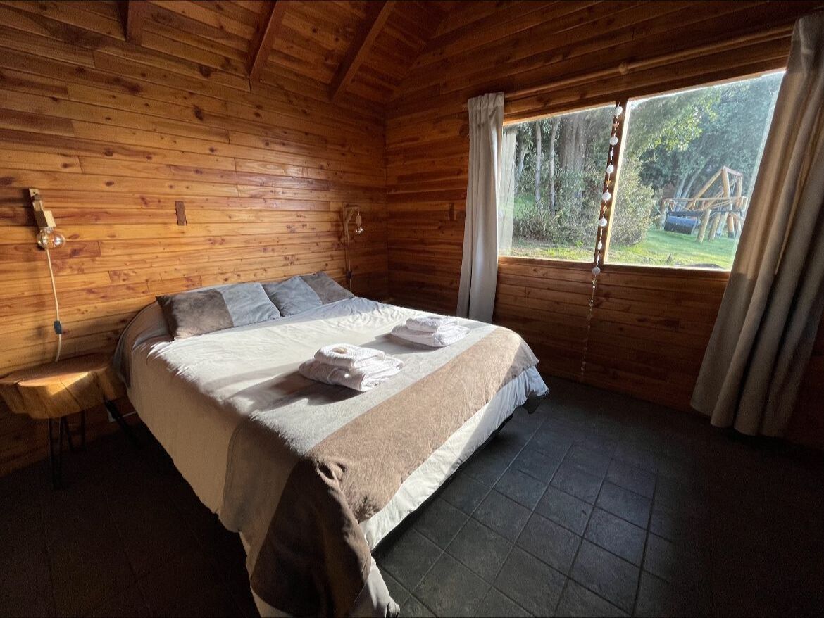 A wooden cabin bedroom with a bed, a nightstand, and a window with curtains.