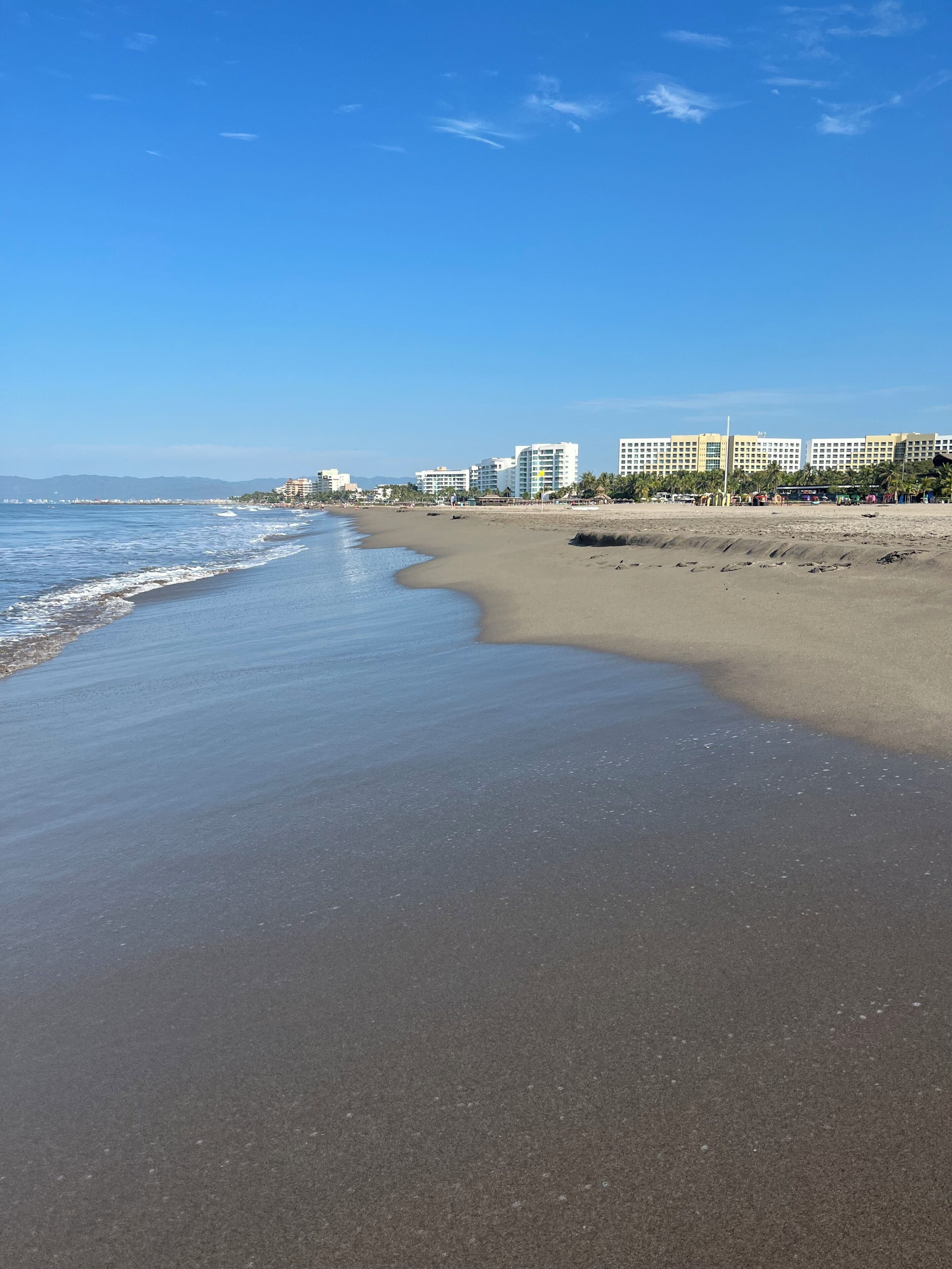 A beach with a clear sky, a sandy shore, and a row of multi-story buildings in the background.
