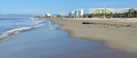 A beach with a clear sky, a sandy shore, and a row of multi-story buildings in the background.