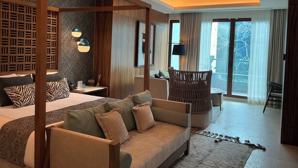 A modern hotel room with a large bed, a sofa, a wooden partition, and a view of the outdoors.