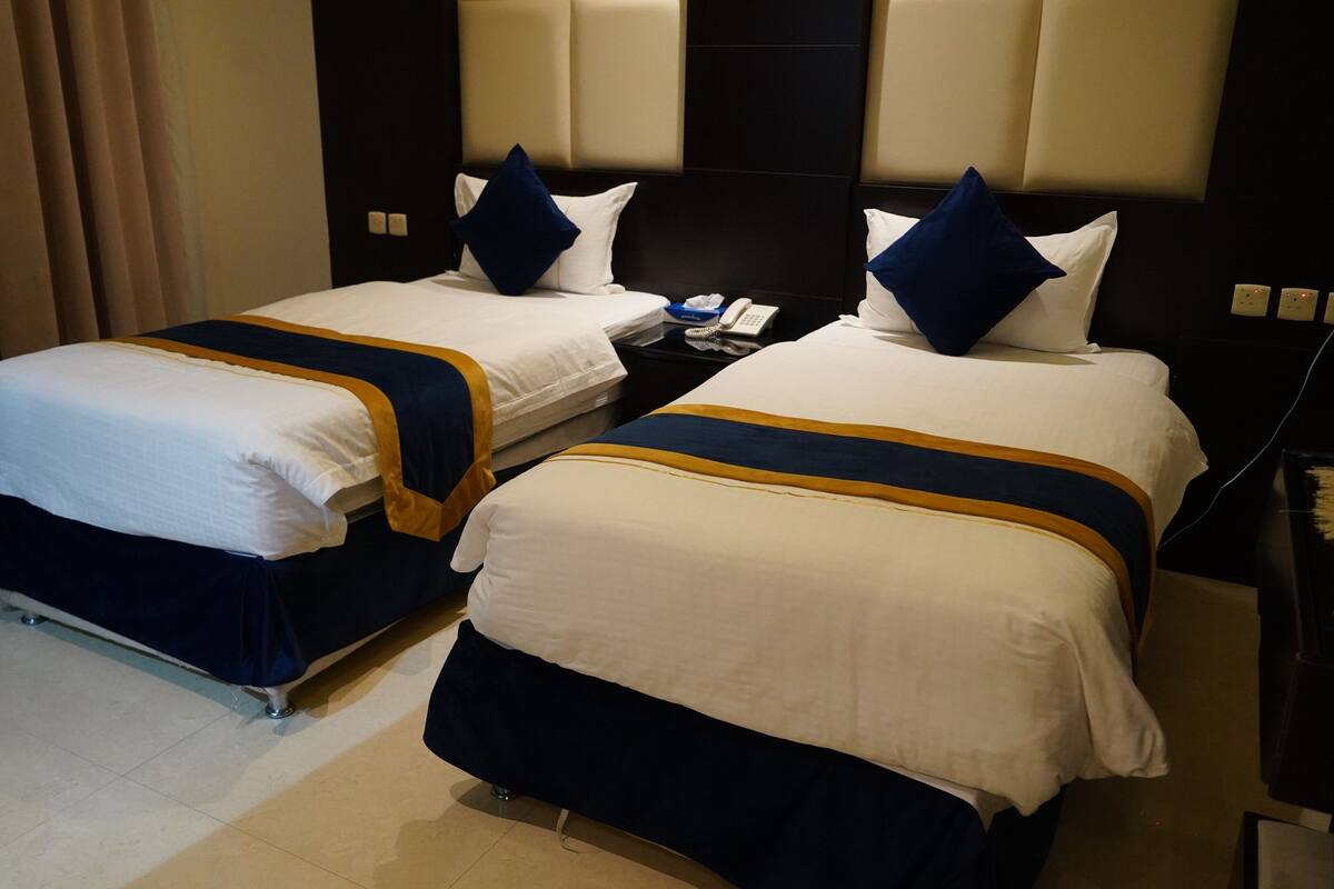 A hotel room with two beds, each with white bedding and blue accents. There is a nightstand with a telephone and a lamp between the beds.