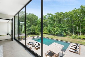 A modern outdoor pool area with lounge chairs and a table, surrounded by glass walls and a view of a forested area. - Glass Villa East Hampton (East Hampton)
