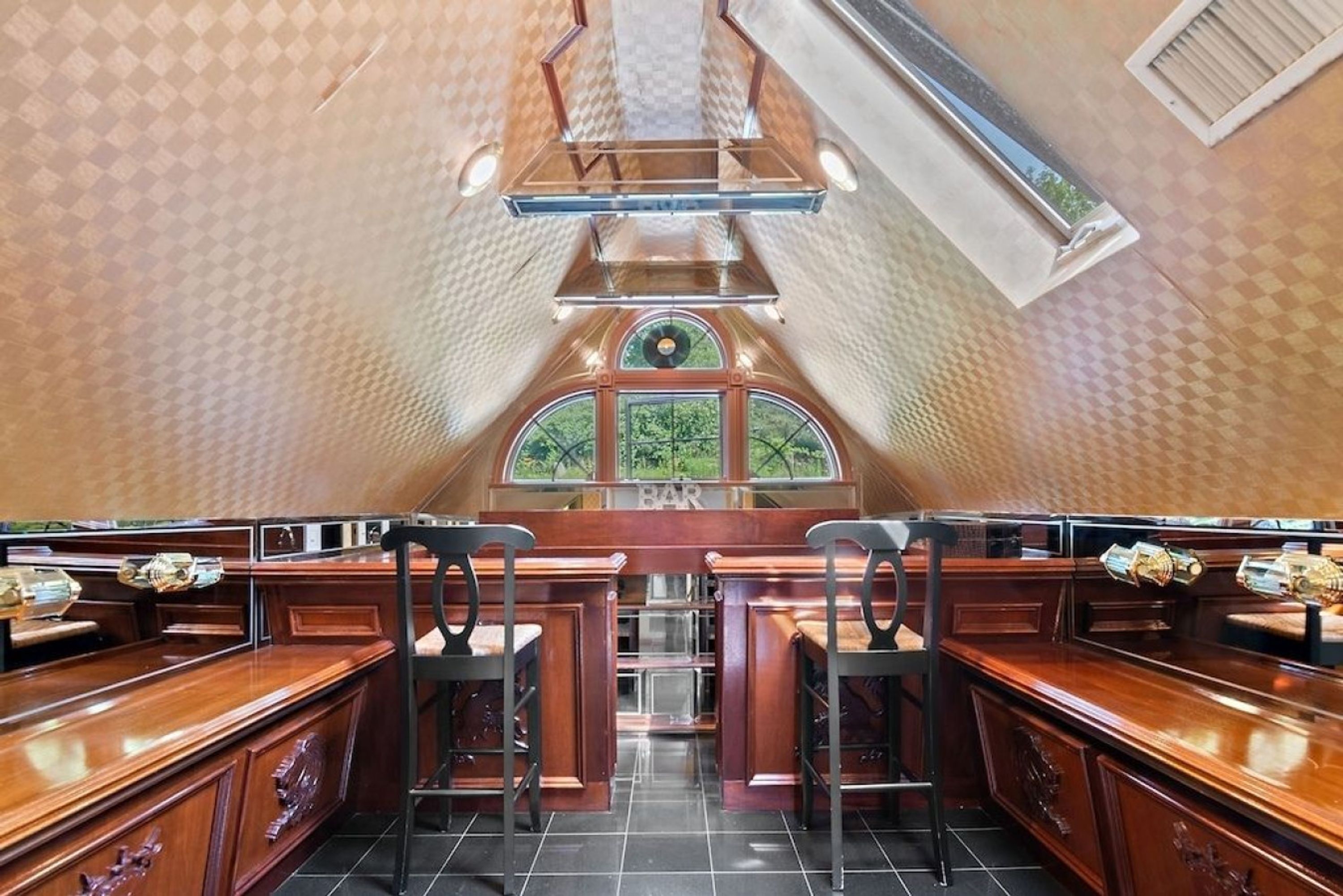 A bar area with wooden bar counters and stools, a large window, and a ceiling with a geometric pattern.