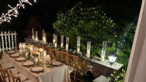 Een buitenterras met een lange tafel gedekt voor een maaltijd, omringd door hoge kaarsen en lichtslingers.