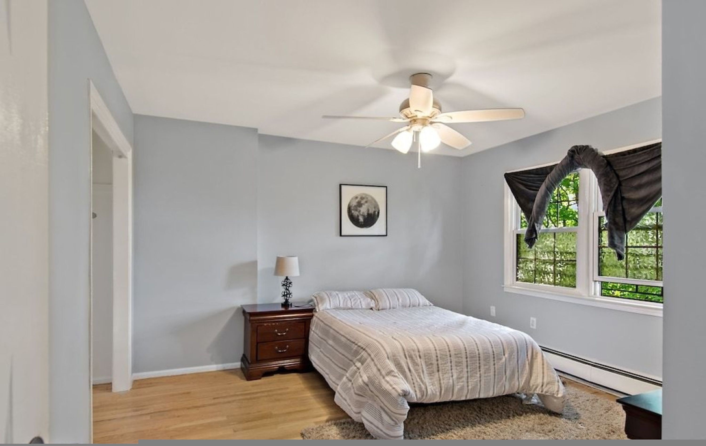 A bedroom with a bed, a nightstand, a ceiling fan, a window with curtains, and a framed picture on the wall.