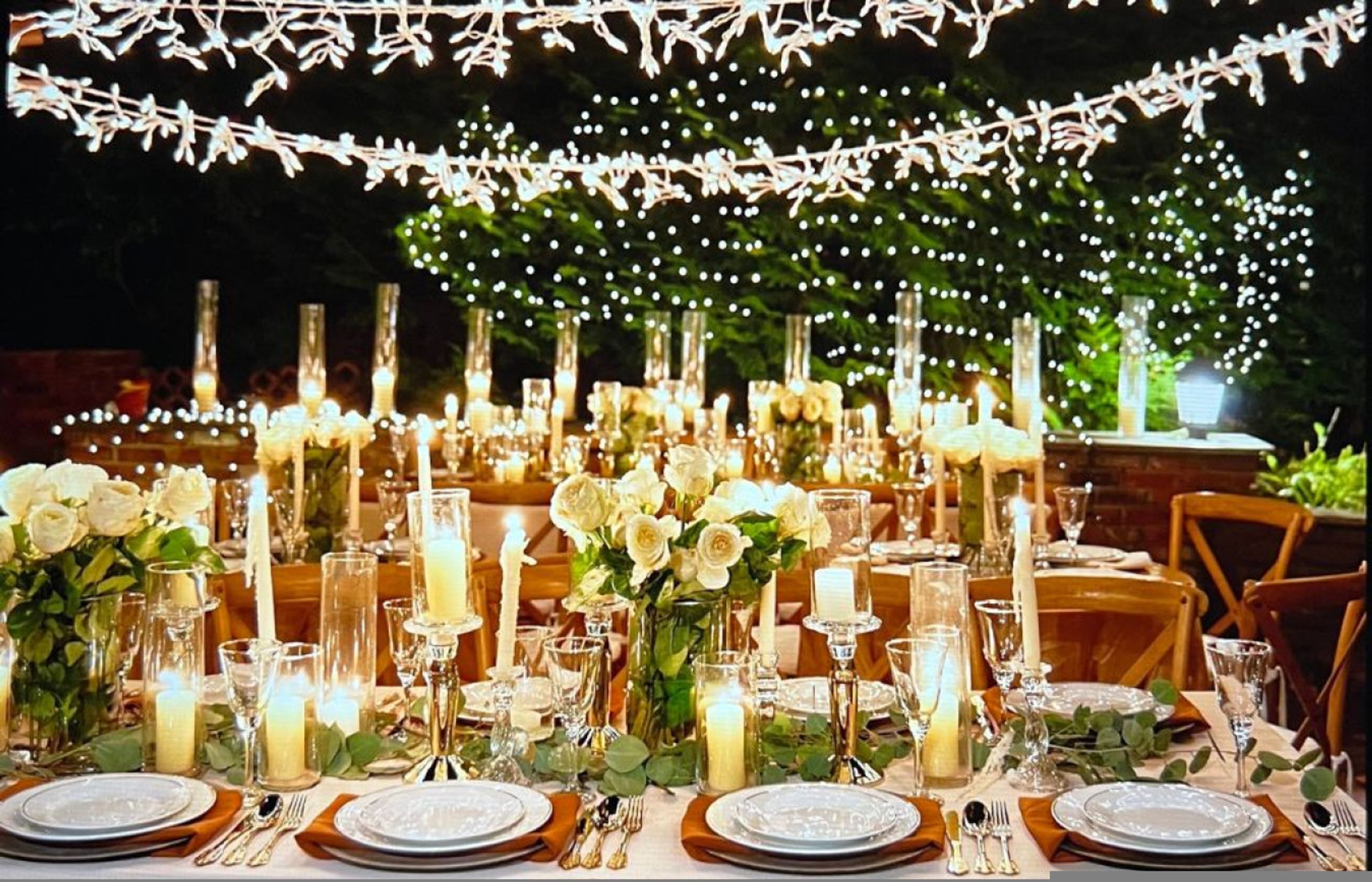 A banquet table set with white roses, candles, and glassware, decorated with string lights.