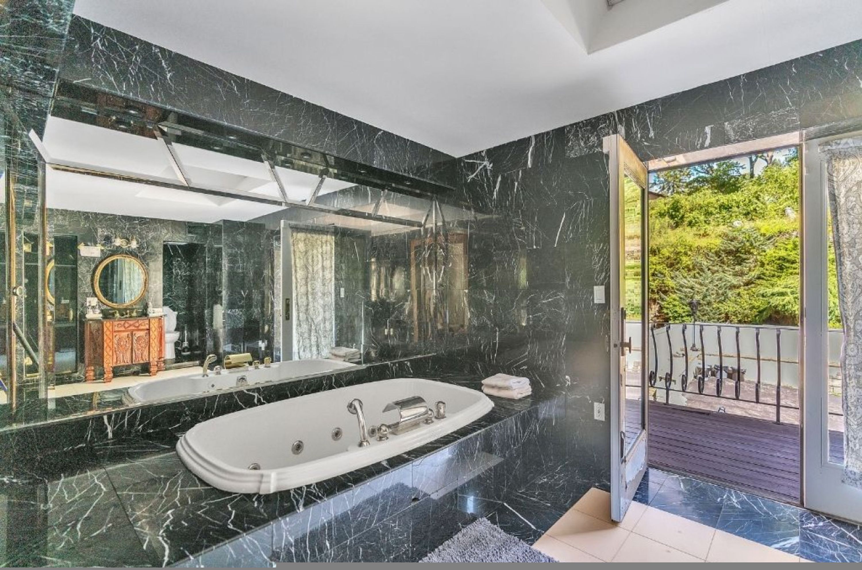 Un baño moderno con una bañera independiente grande, un tocador con espejo y vistas a un balcón con vegetación.