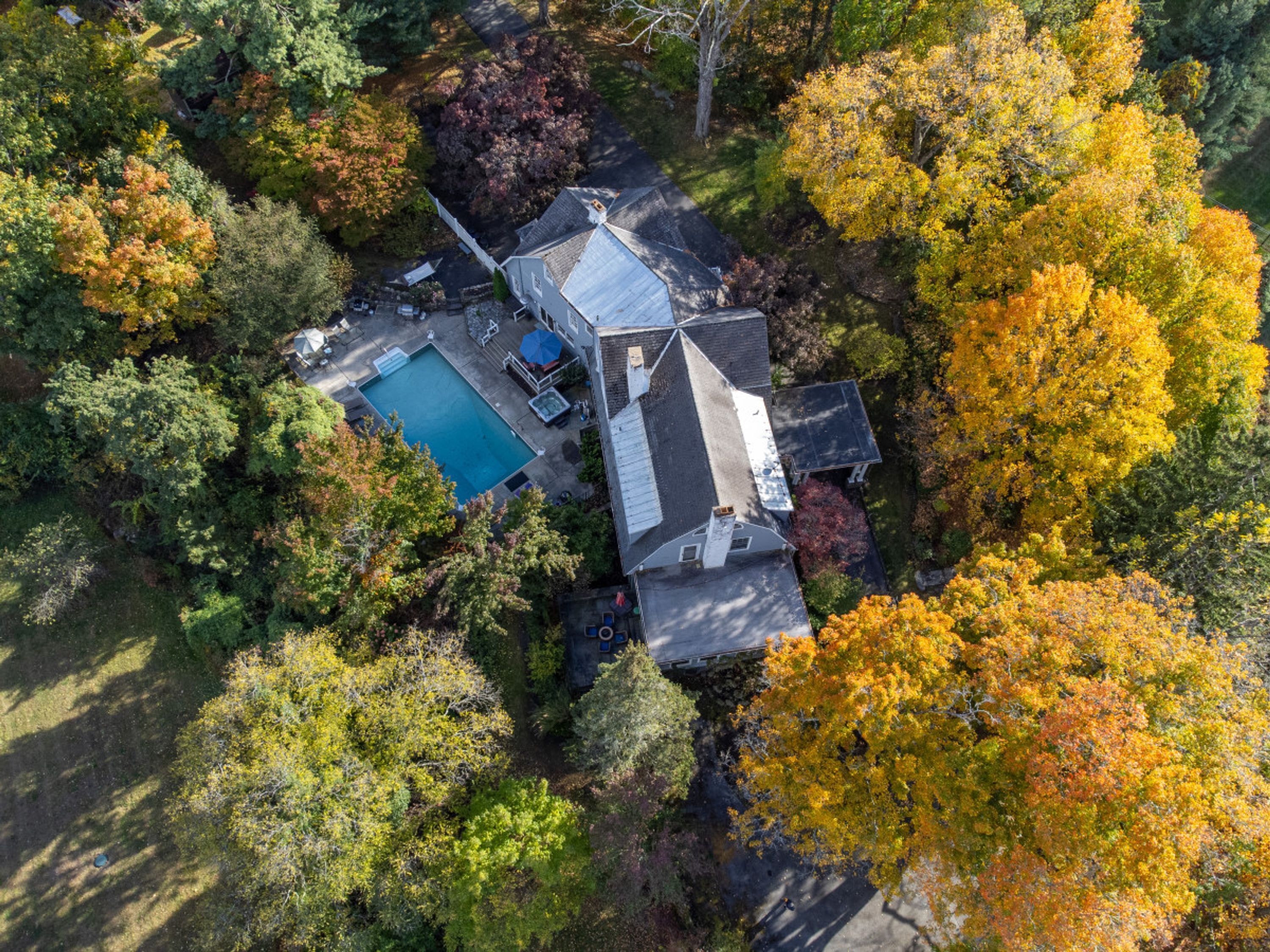 An aerial view of a house with a swimming pool, surrounded by dense trees with autumn foliage.
