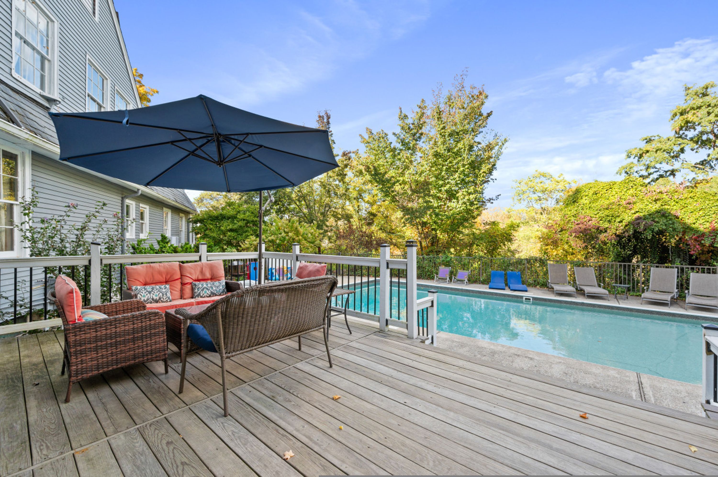 Una terraza de madera con mobiliario exterior, una piscina y vistas a árboles y una casa.