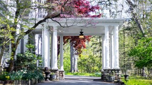 Une allée couverte, ornée de colonnes blanches, entourée d’une végétation luxuriante et d’arbres aux feuilles d’automne.