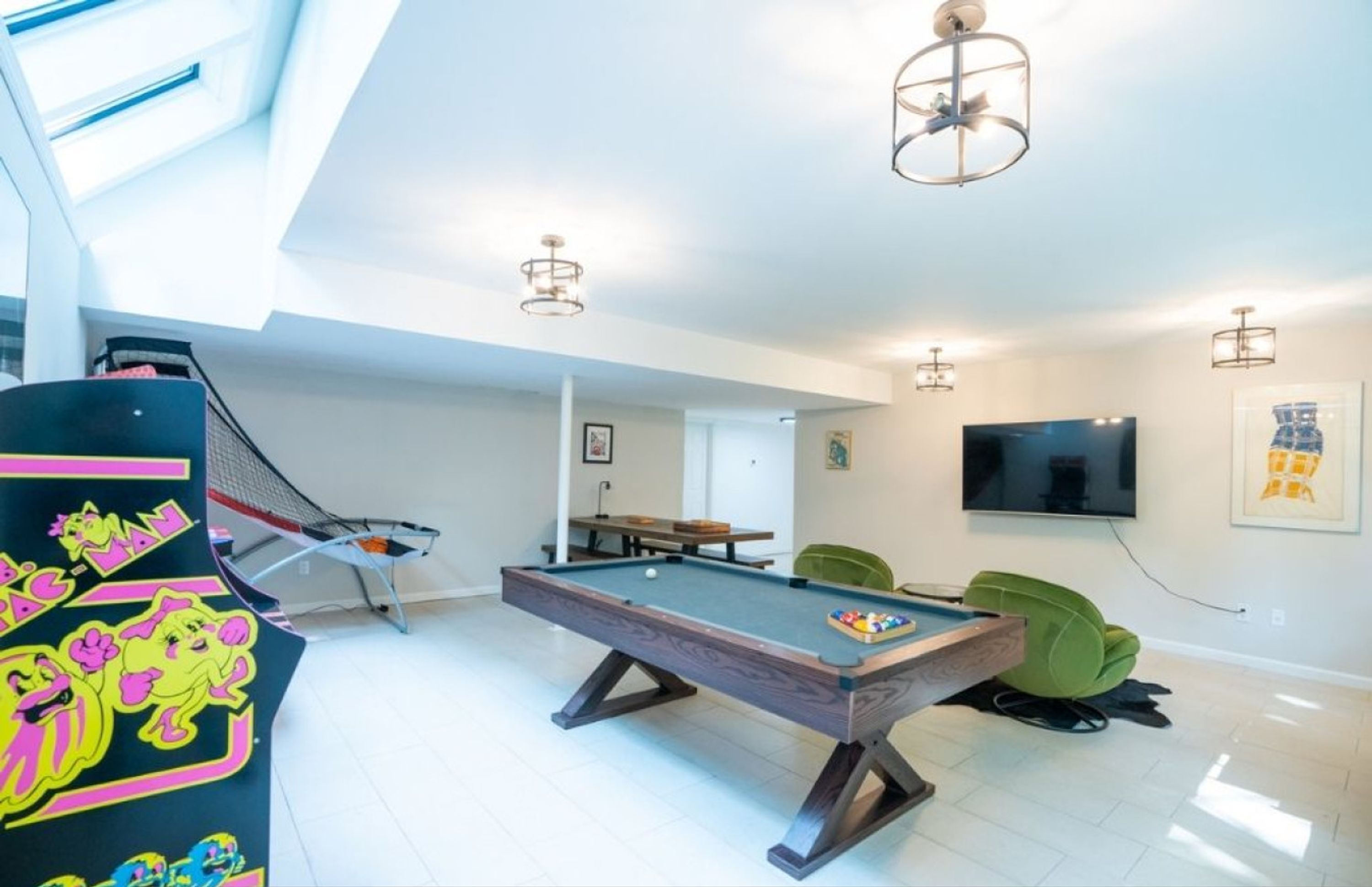 Une pièce spacieuse équipée d’une table de billard, d’une table de ping-pong et d’une table de jeu de fléchettes. Un téléviseur est fixé au mur et une chaise verte se trouve près de la table de billard.