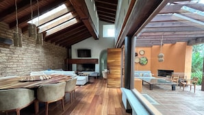 Uma sala de estar ampla com uma mesa de jantar de madeira, assentos confortáveis, lareira e uma grande janela com vista para um jardim.