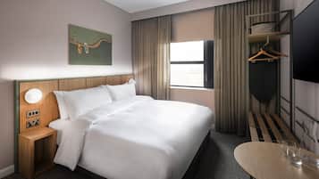 Ein Hotelzimmer mit einem großen Bett, einem Nachttisch mit Lampe, einem an der Wand befestigten Fernseher, einem Kleiderständer und einem kleinen Tisch mit Gläsern.