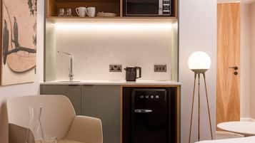 Uma cozinha compacta equipada com micro-ondas, cafeteira e frigorífico. Inclui uma cadeira e uma pequena mesa com copos.