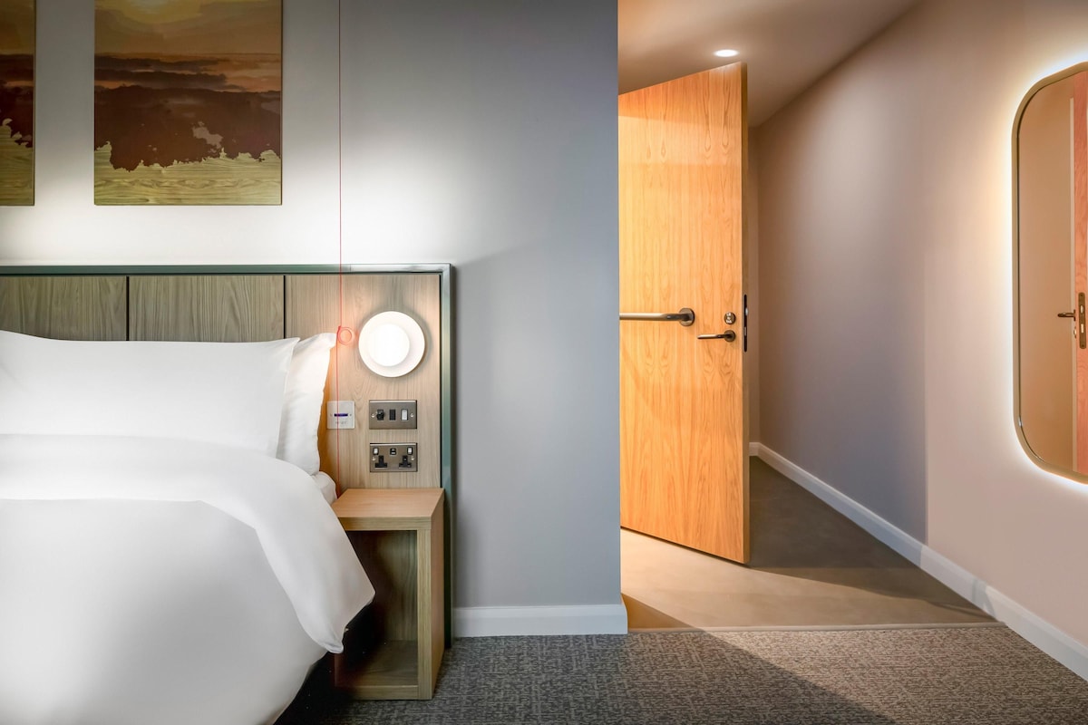 Ein Hotelzimmer mit Bett, Nachttisch, Wandlampe und einer Tür zu einem anderen Raum.