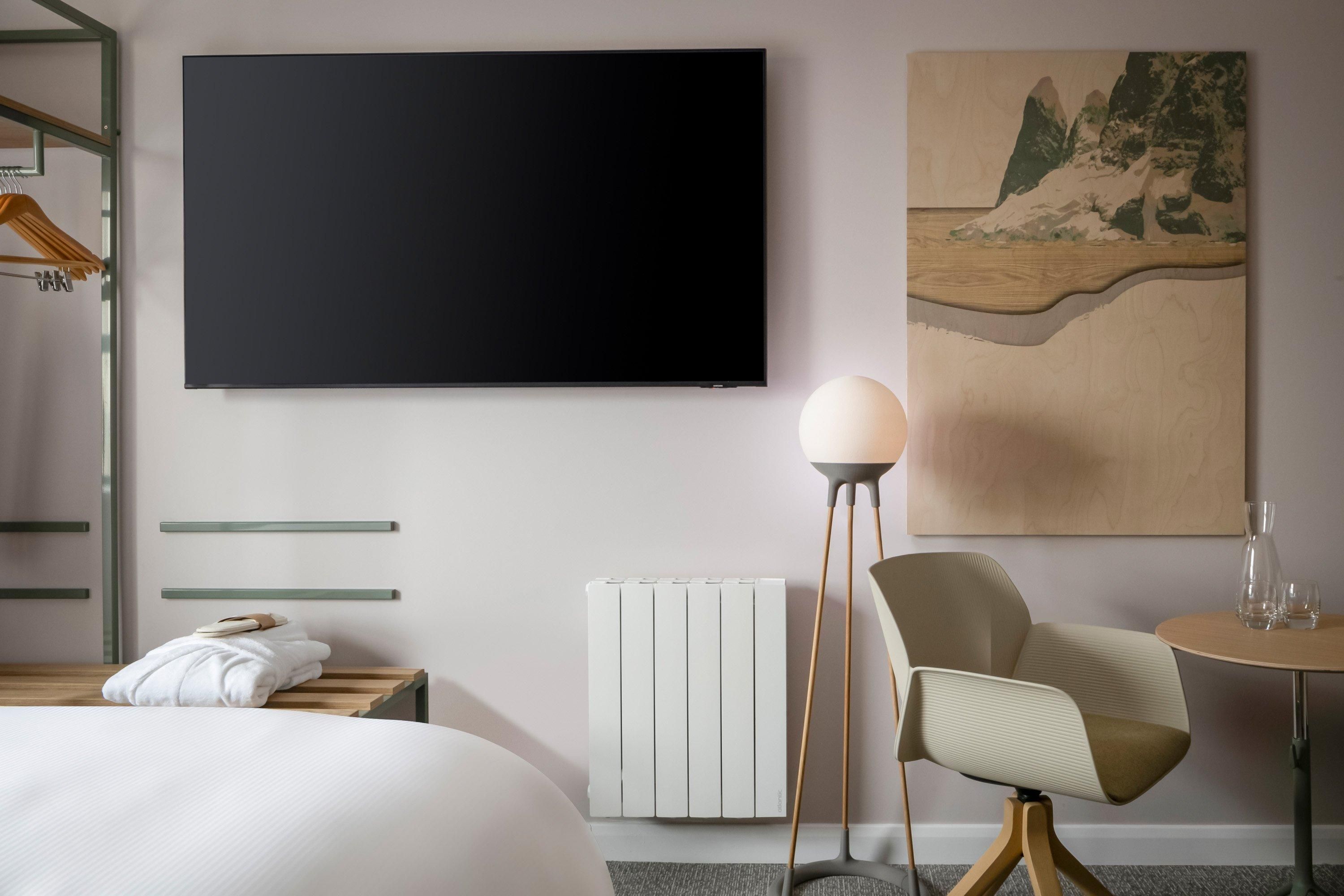 A hotel room with a bed, a flat-screen TV, a radiator, a chair, a table, and a painting on the wall.