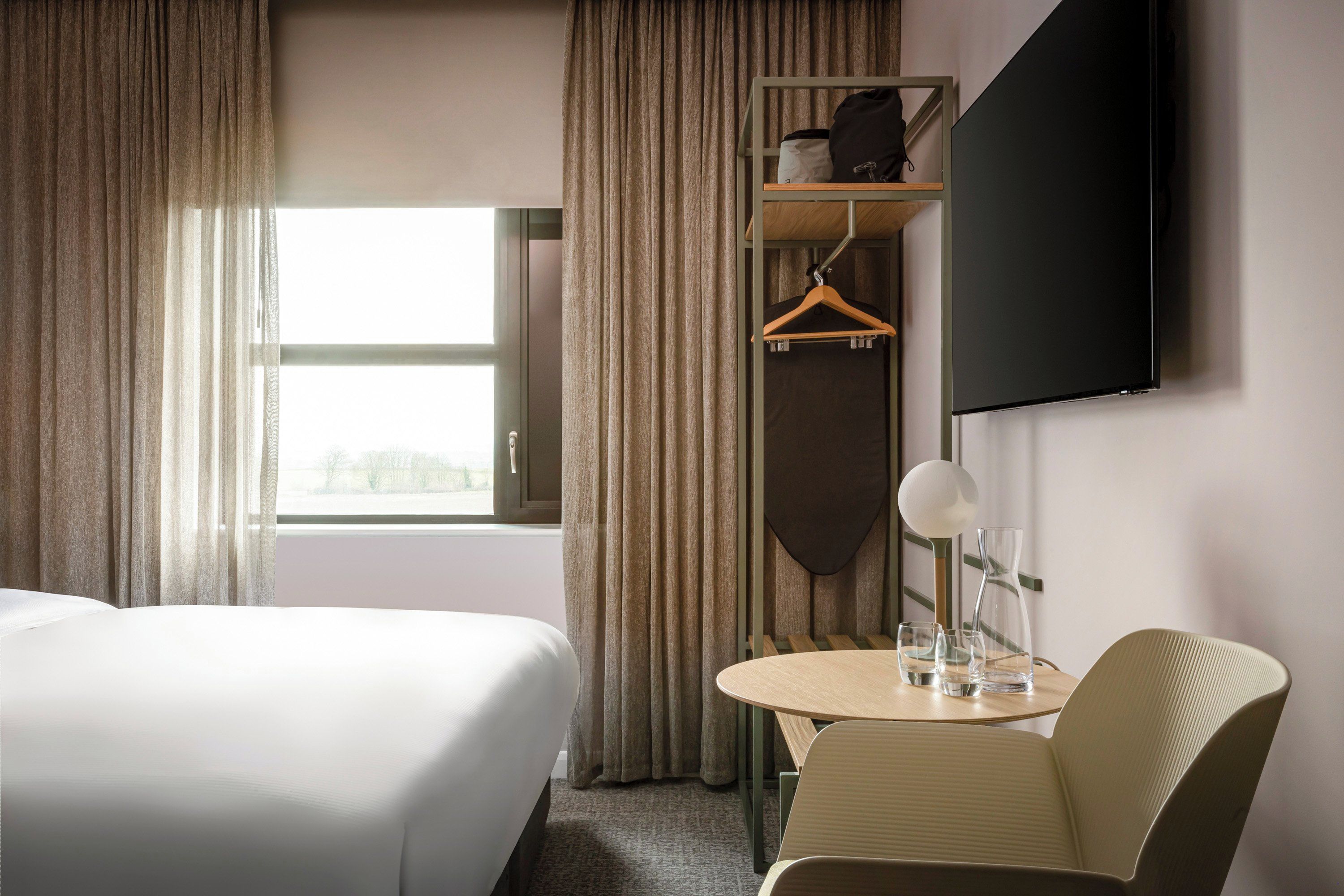 Una habitación de hotel con dos camas, una mesita con vasos, un perchero y un televisor montado en la pared.