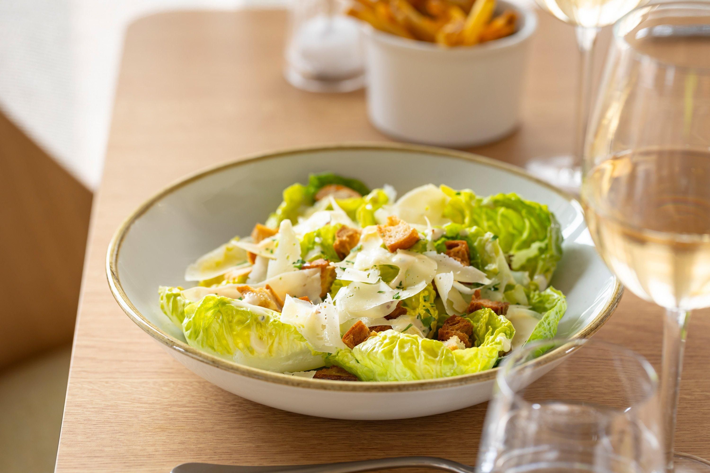 Ensalada César con crutones y queso, acompañada de una copa de vino blanco sobre una mesa de madera.