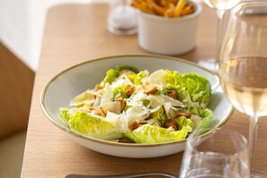 A plate of Caesar salad with croutons and cheese, accompanied by a glass of white wine on a wooden table. - Voco Zeal Exeter Science Park (Exeter)