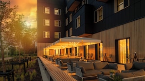 Un área de lounge moderna al aire libre con asientos y sombrillas, junto a un edificio con varias ventanas y balcones.