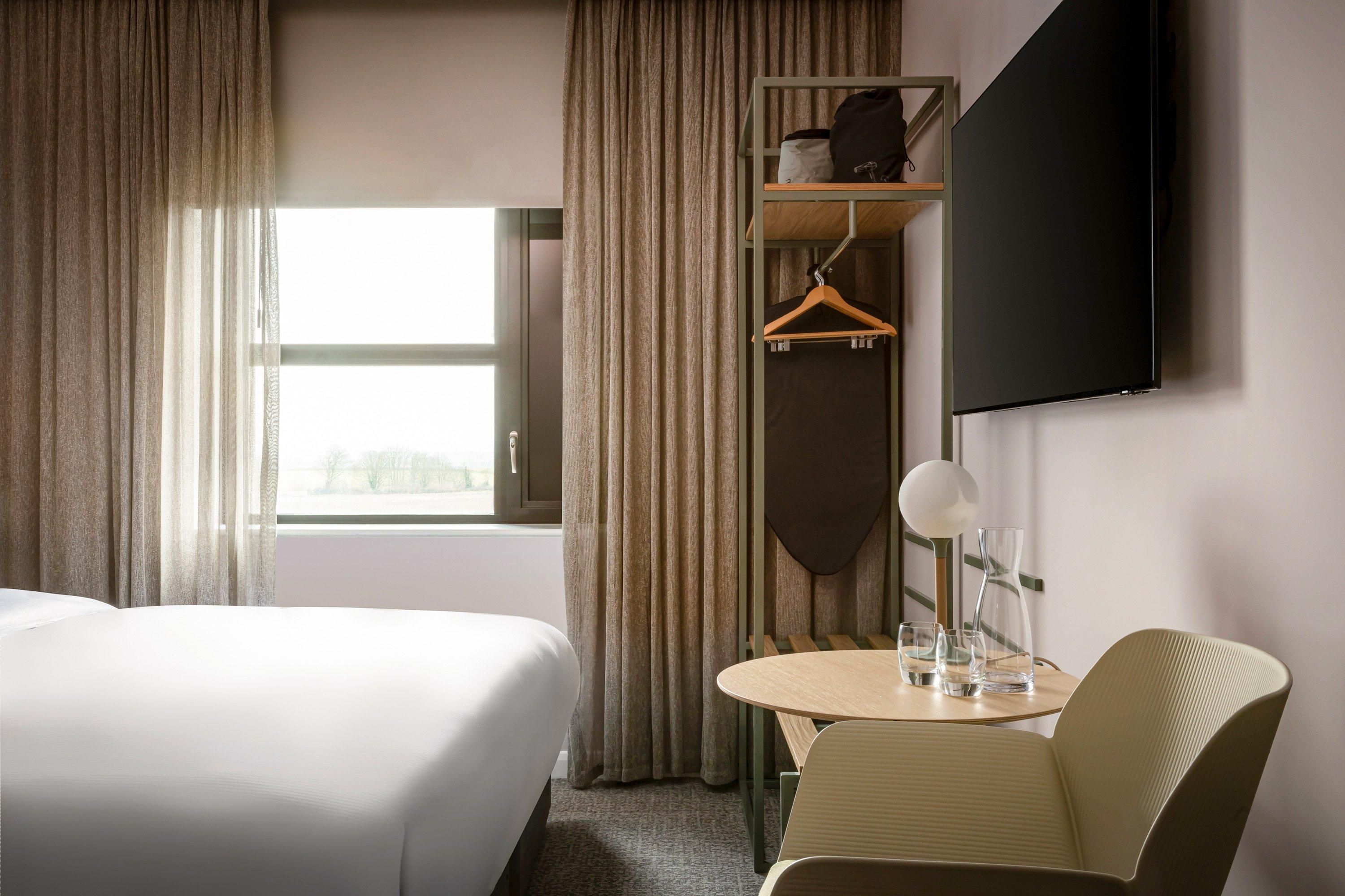Habitación de hotel con cama, escritorio con silla, televisor montado en la pared y ventana con cortinas.