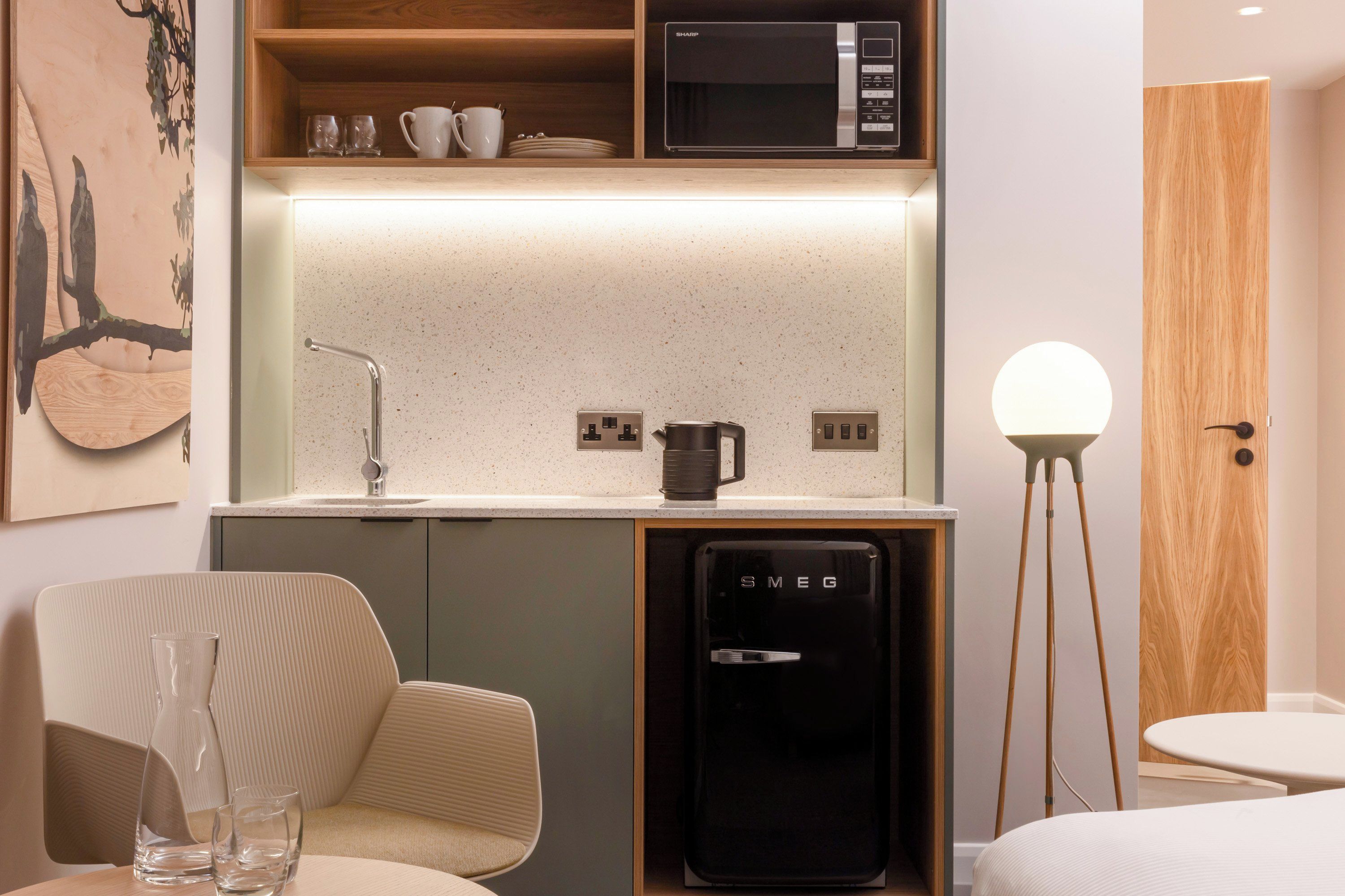 Eine moderne Küchenzeile mit Mikrowelle, Kaffeemaschine und Kühlschrank. Es gibt eine Essecke mit einem Stuhl und Tisch sowie ein Bild an der Wand.