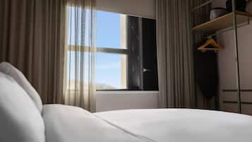 Una habitación de hotel con cama, ventana con cortinas, estante con una prenda colgada y vista al cielo.