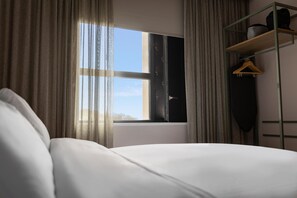 A hotel room with a bed, a window with curtains, a shelf with a hanging garment, and a view of the sky. - Voco Zeal Exeter Science Park (Exeter)