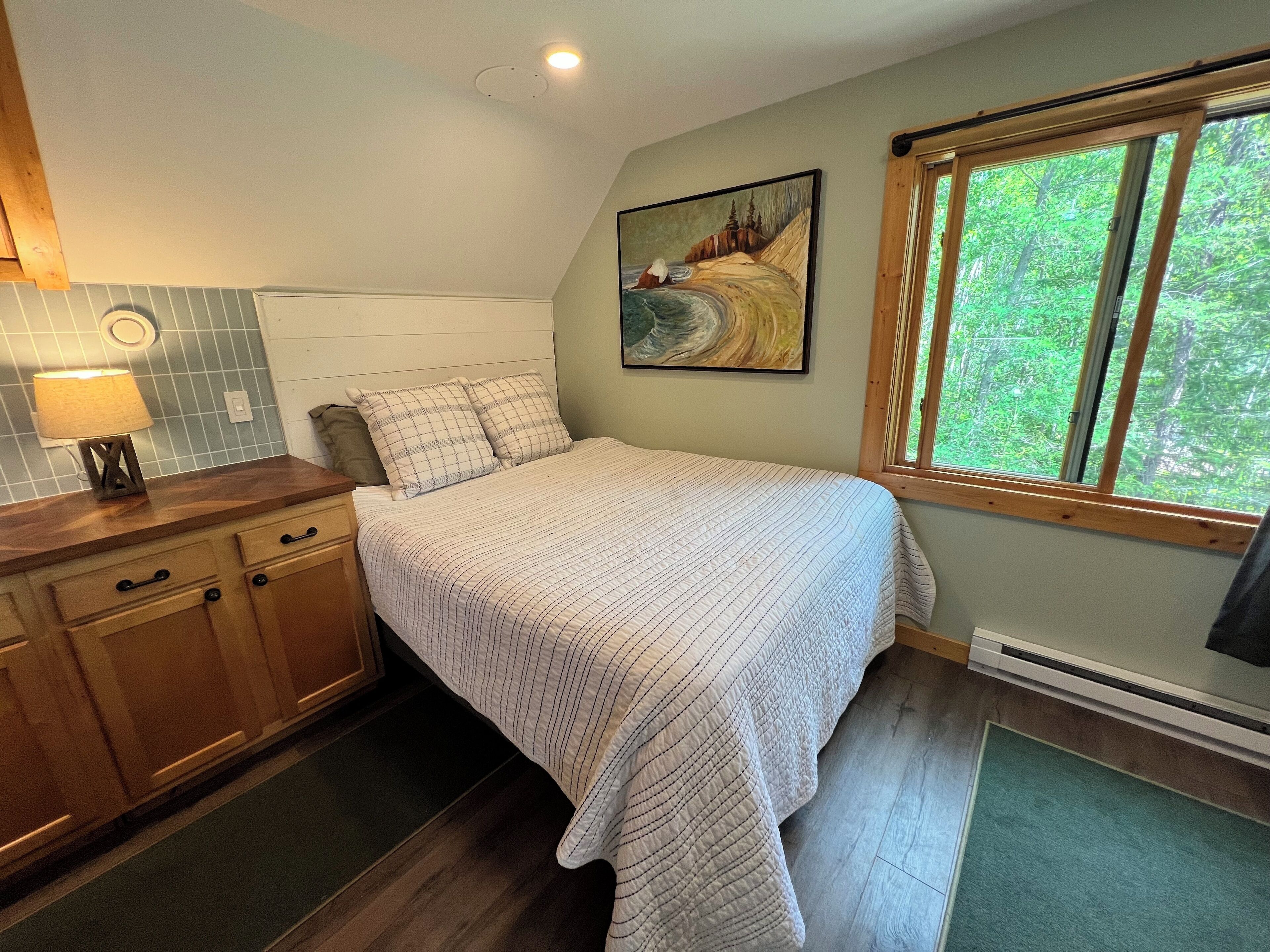 A bedroom with a bed, a nightstand, a lamp, a painting, and a window with a view of trees.