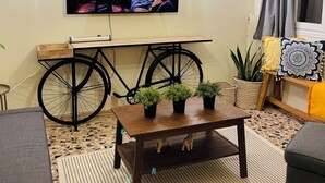Un salon chaleureux avec un téléviseur fixé au mur, une table en bois agrémentée de plantes en pot, une roue de vélo servant de table d’appoint et un tapis à motifs.