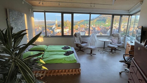 Una habitación moderna con un ventanal grande, una cama, dos sillas, una mesita y un televisor.