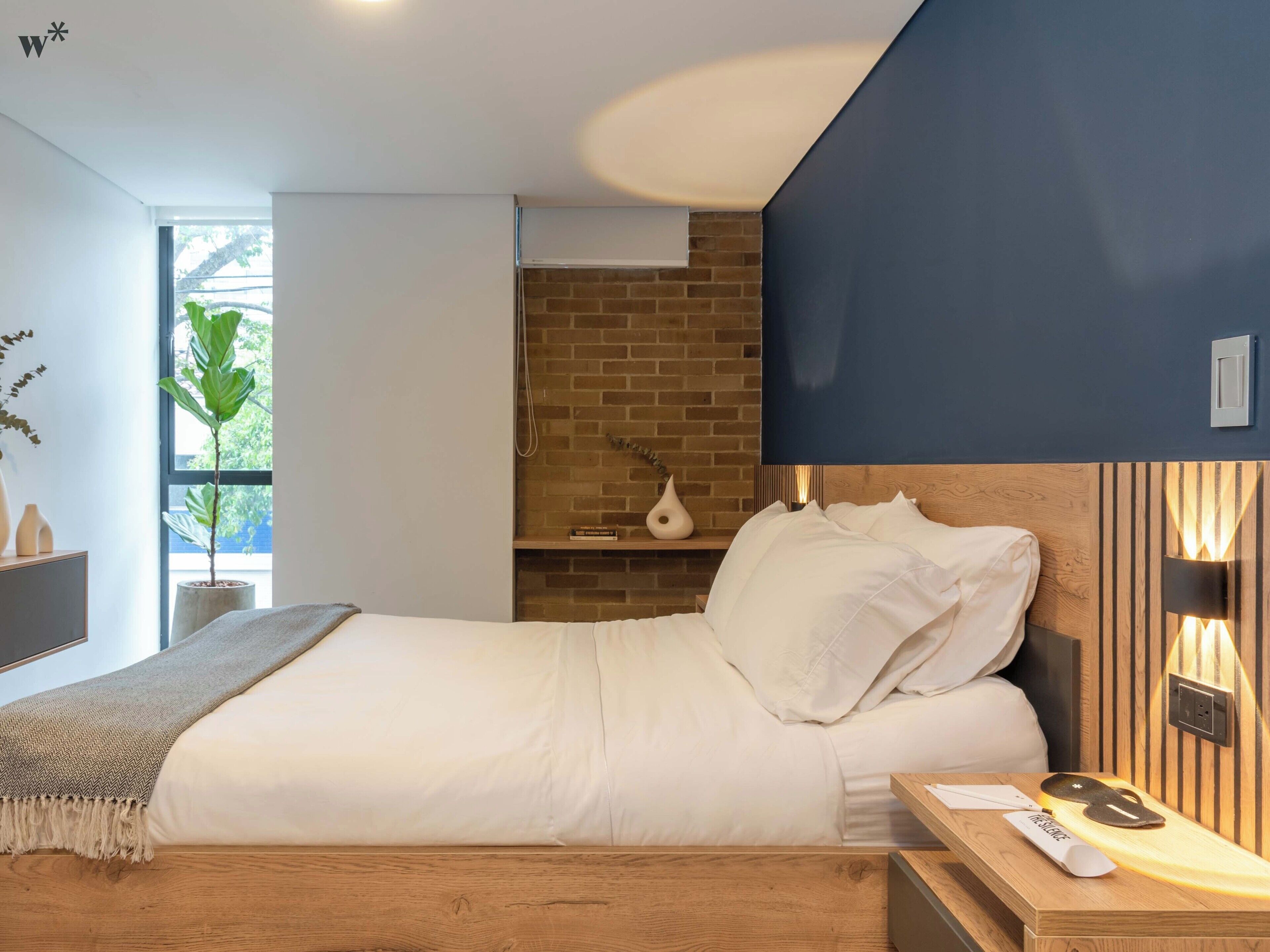 A modern hotel room with a large bed, a wooden nightstand, a brick accent wall, and a large window with a plant.