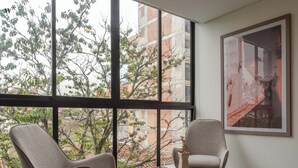 A modern room with two chairs facing a large window, a round table with a vase and book, and a framed picture on the wall. - Ganesh by Wynwood House (Medellín)