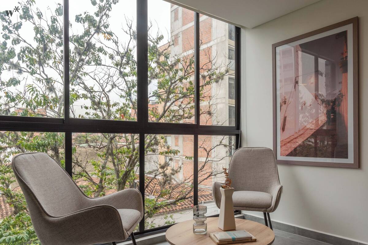 A modern room with two chairs facing a large window, a round table with a vase and book, and a framed picture on the wall.