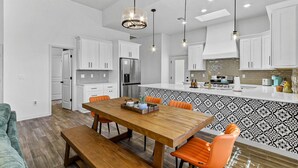 A modern kitchen with white cabinets, a wooden dining table, and orange chairs. - Book a Winter Stay! Hot Tub & Brand New Modern Cozy Home (Las Cruces)