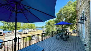 Eine Holzterrasse mit Picknicktischen und Sonnenschirmen, mit Blick auf einen Parkplatz mit Wohnmobilen und einen See.