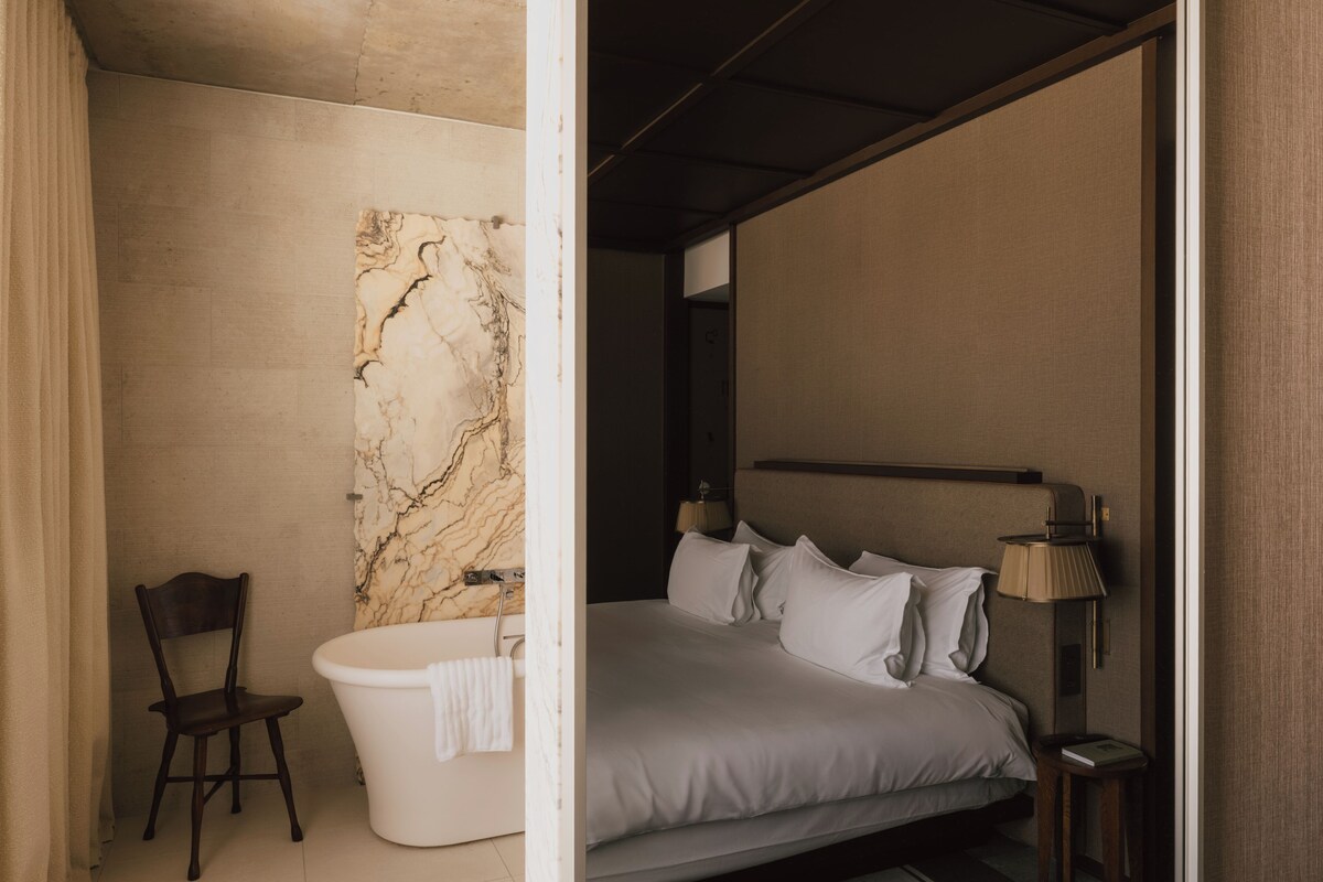 A modern bedroom with a large bed, a marble wall, a chair, and a bathtub.