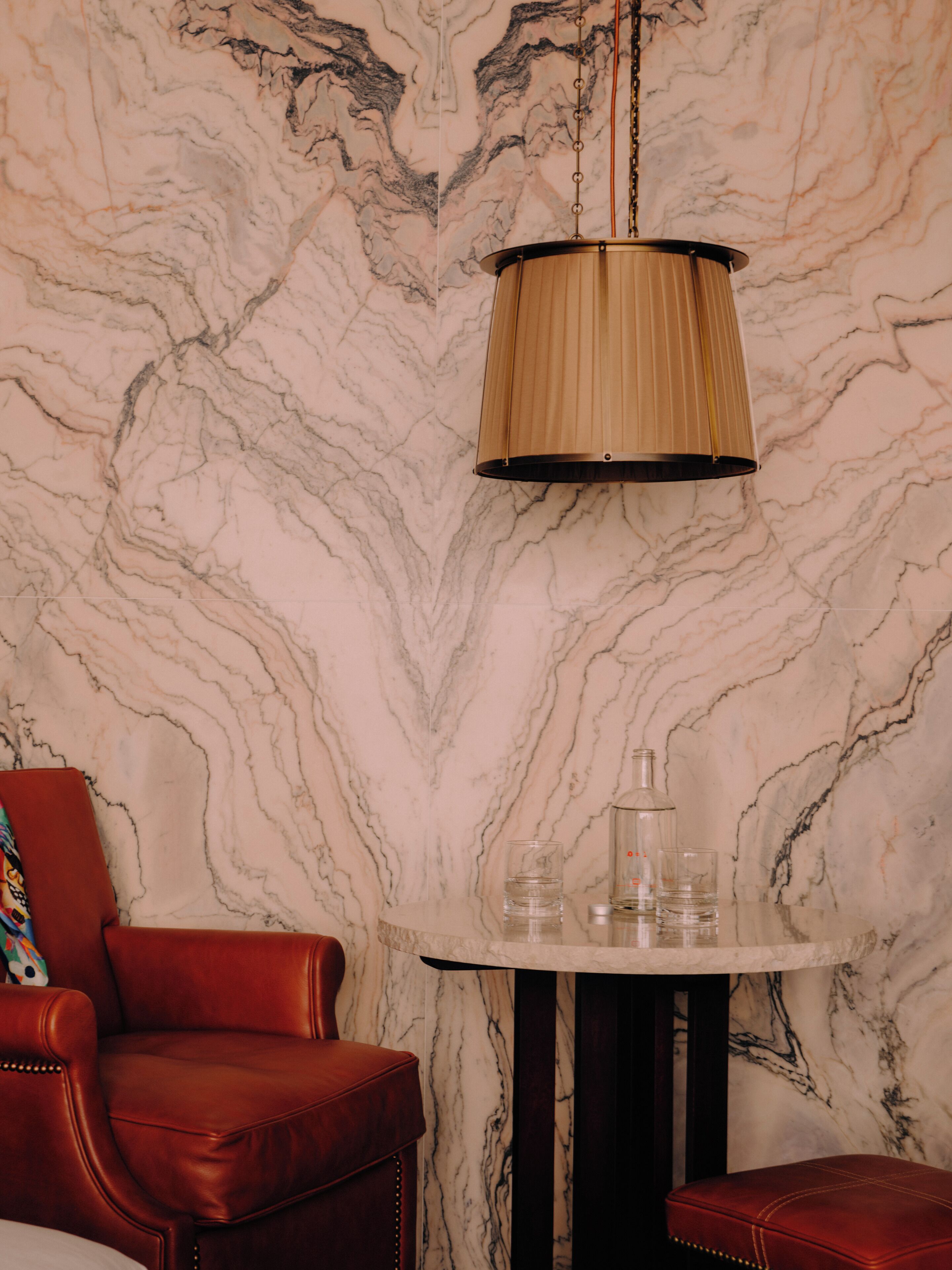 A room with a marble wall, a wooden chair, a table with glasses and a bottle, and a hanging lamp.