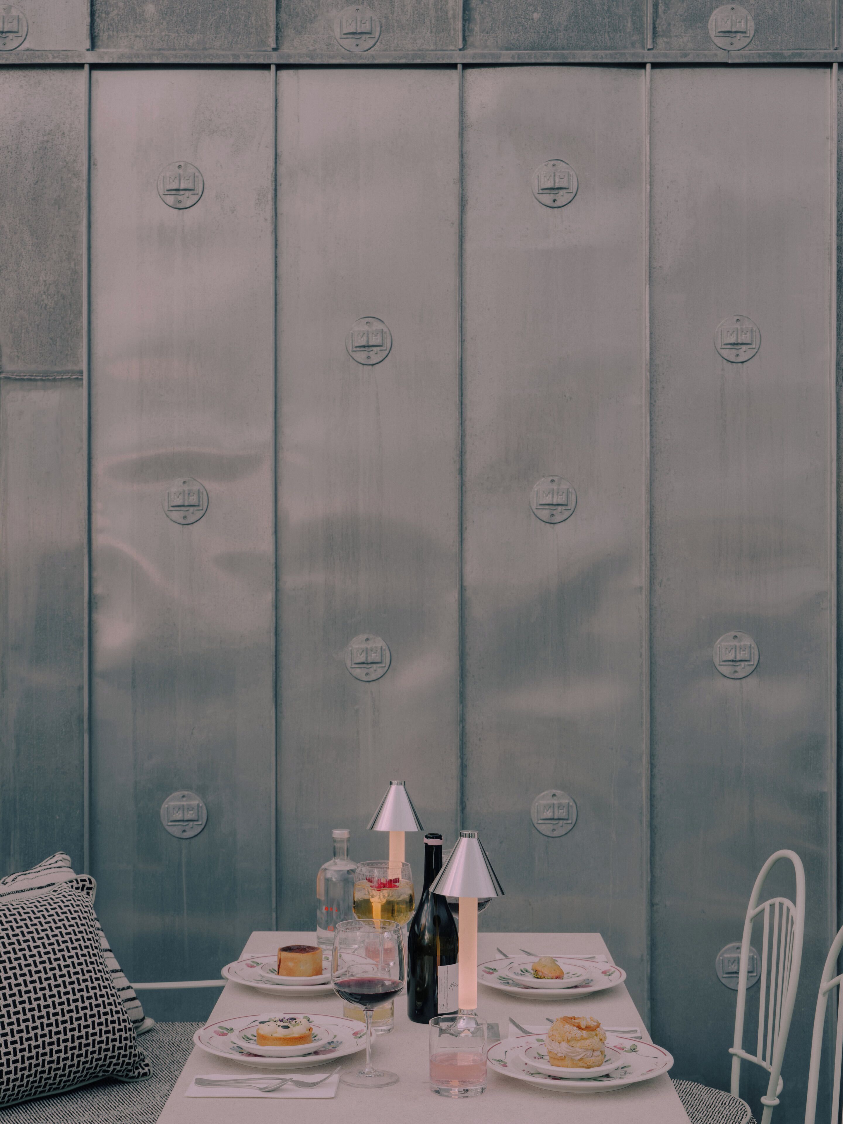 A table set with plates, glasses, and bottles, against a metallic wall with circular patterns.