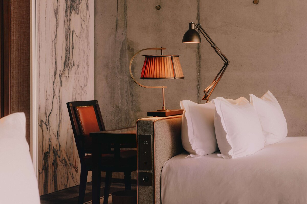 A hotel room with a bed, a desk, a chair, and a lamp.
