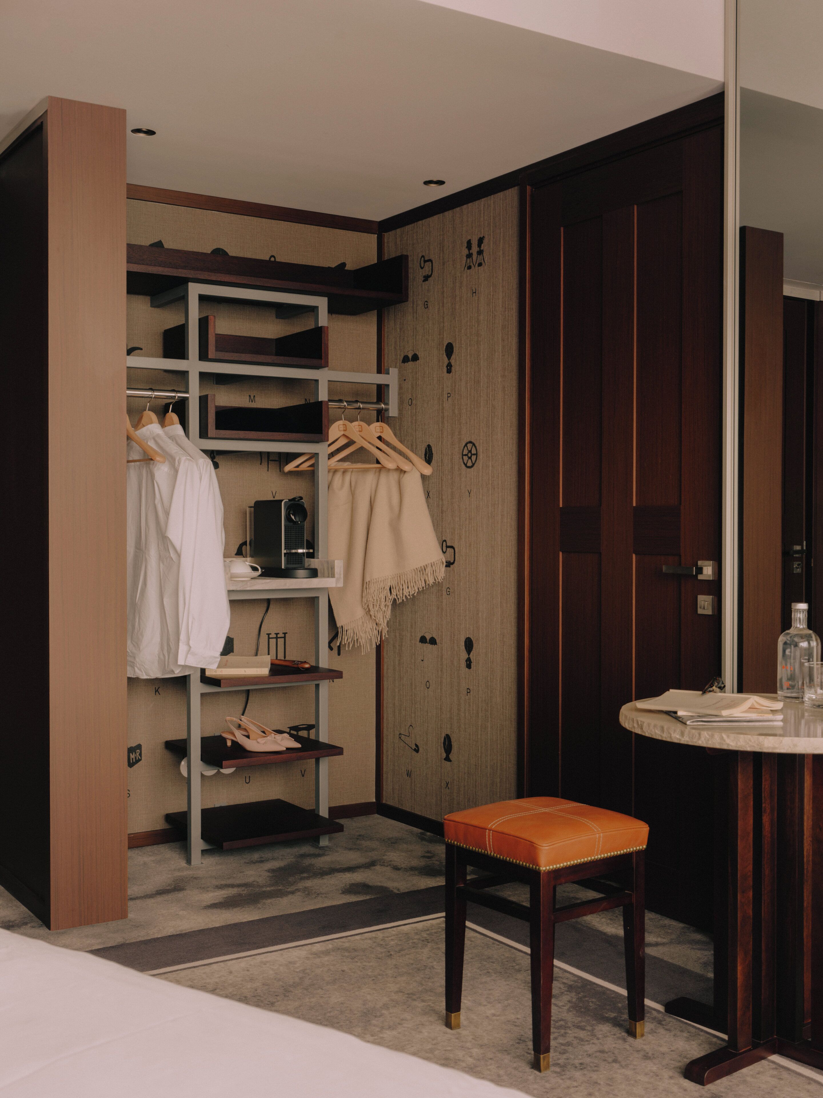 A compact bedroom with a wooden wardrobe, a small table with a chair, and a closet with hanging clothes.