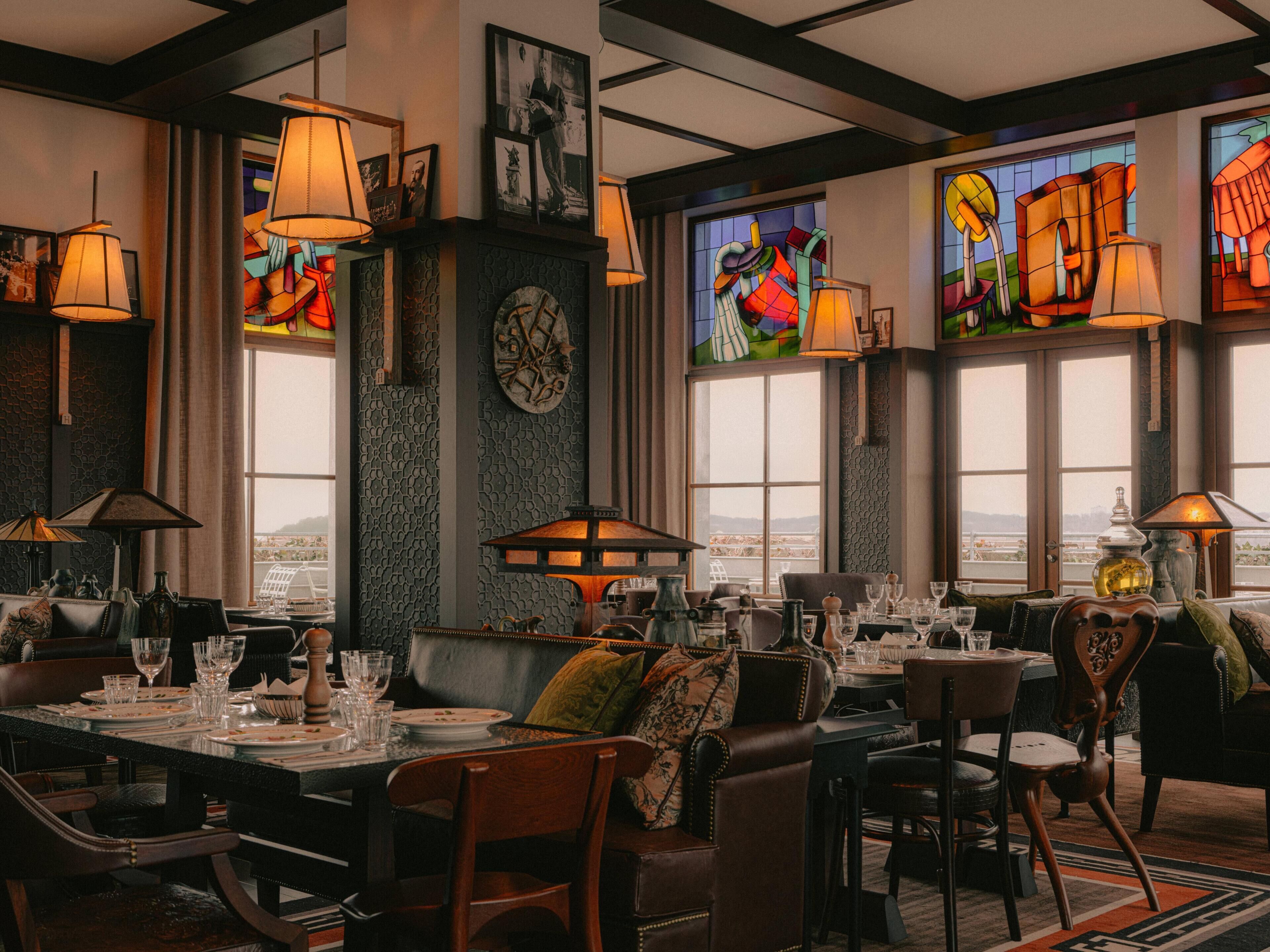 A restaurant interior with high ceilings, large windows, and multiple dining tables set with glasses and plates.