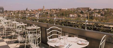 An outdoor dining area with tables and chairs, overlooking a cityscape with buildings and a tower.