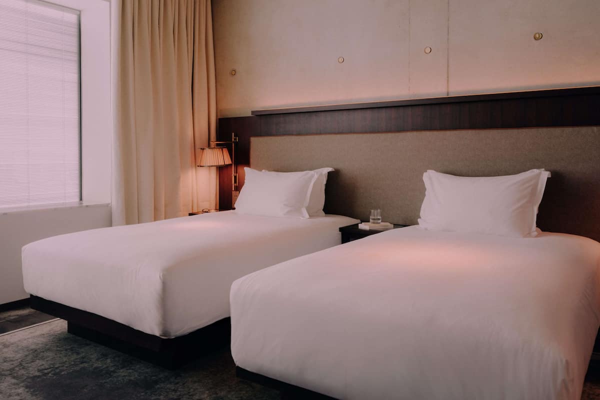 A hotel room with two beds, a window with blinds, a lamp, and a nightstand with a glass of water.