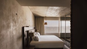 A modern hotel room with a large bed, a nightstand, a lamp, and a view of the city through large windows. - Maison Heler Metz, Curio Collection By Hilton (Metz)