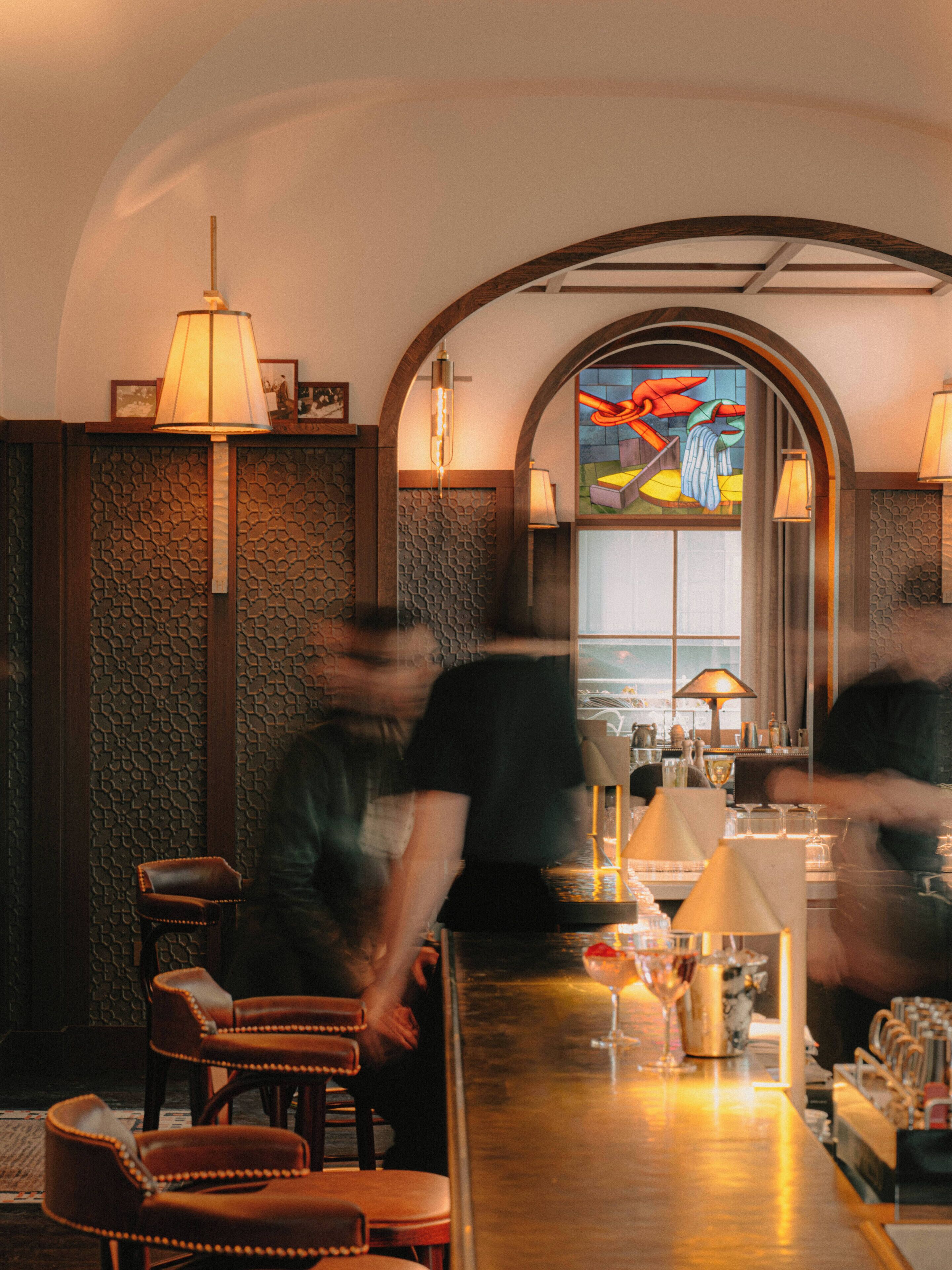 A bar area with a wooden counter, leather chairs, and a stained glass window in the background.