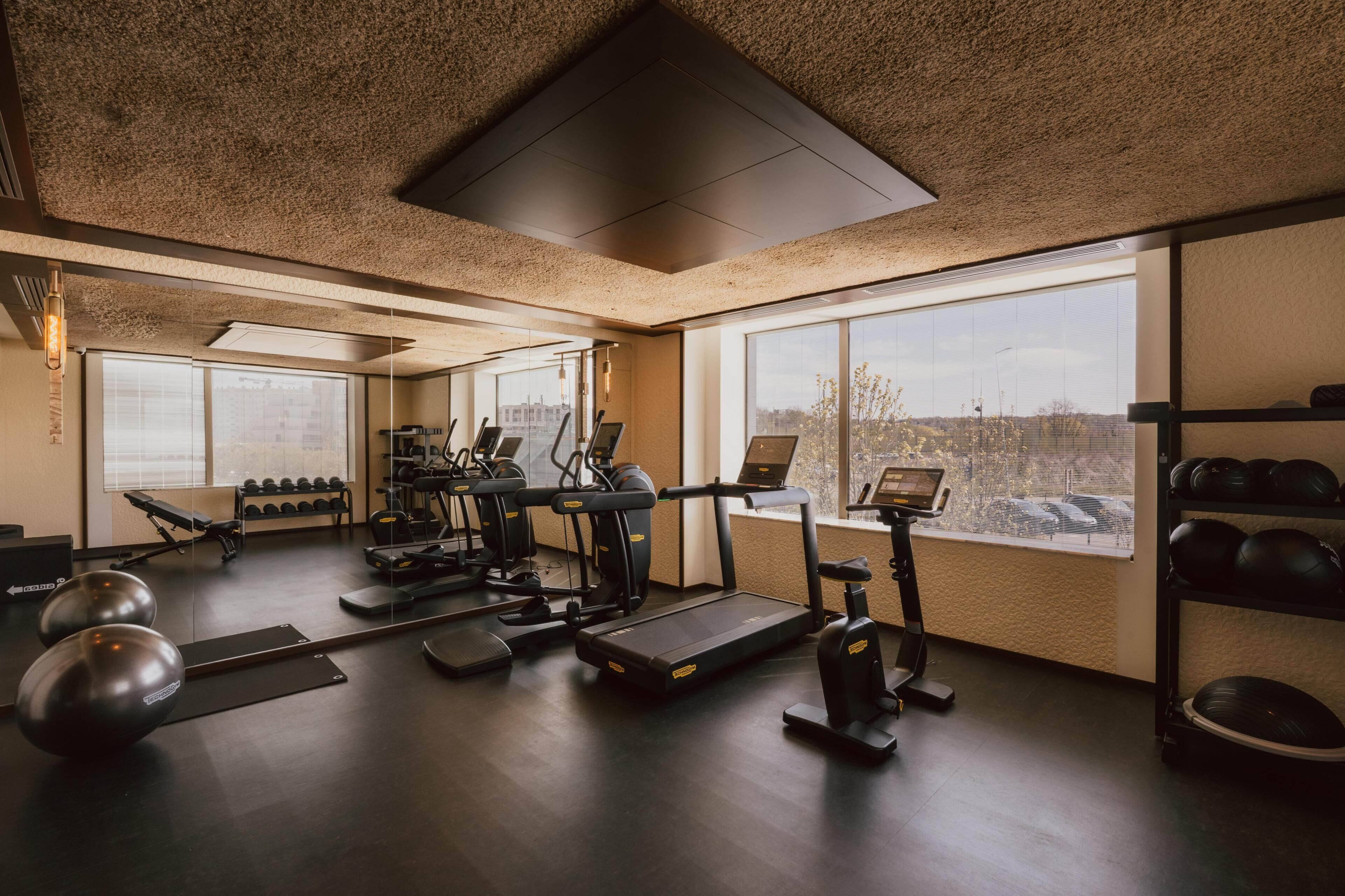 A gym with exercise equipment, a bench, dumbbells, and a large window with a city view.