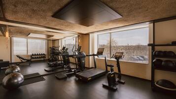 A gym with exercise equipment, a bench, dumbbells, and a large window with a city view.