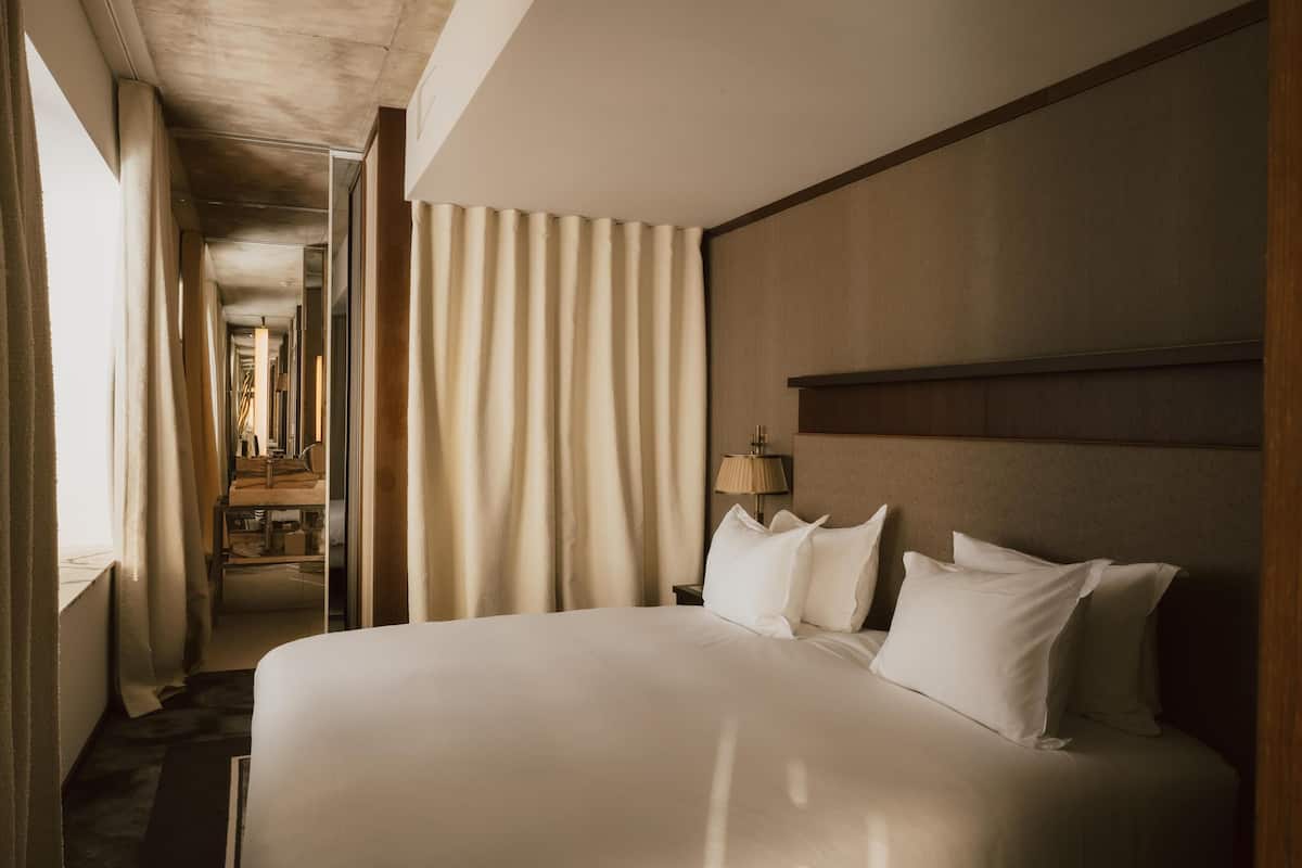 A hotel room with a large bed, a nightstand, a lamp, and a view of a corridor with a mirror.
